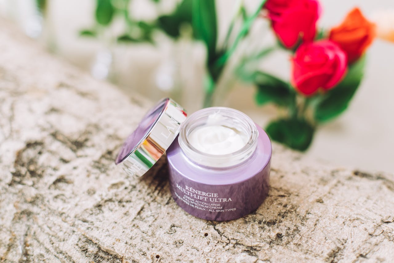 Open jar of luxury skincare cream surrounded by vibrant red roses on a textured surface.
